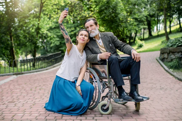 Pretty smiling young woman, care giver or granddaughter in casual hipster clothes, with dreadlock hair, making photo on smartphone with her disabled elderly man, grandfather or patient in wheelchair