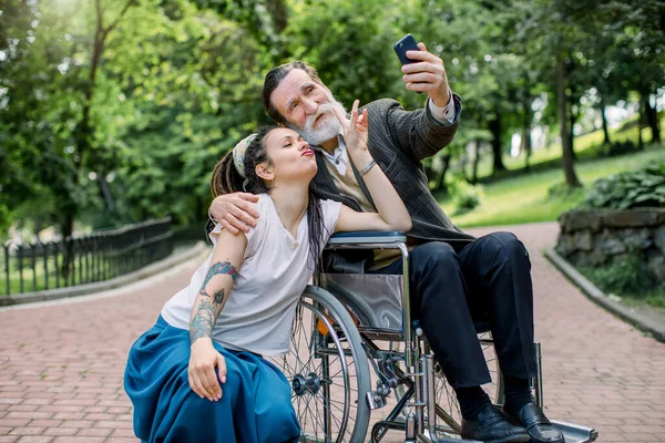 Hipstermädchen mit langen Dreadlocks und gutaussehender bärtiger Großvater im Rollstuhl beim Selfie machen im Park. Lebensstil behinderter Menschen. Pfleger und Patient beim Spaziergang — Stockfoto