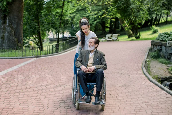 Attraktive junge Hipster-Frau mit langen Dreadlocks, Pflegerin oder Sozialarbeiterin, schiebt Rollstuhl mit ihrem gutaussehenden älteren Mann, der an einem sonnigen Tag im Park spazieren geht — Stockfoto