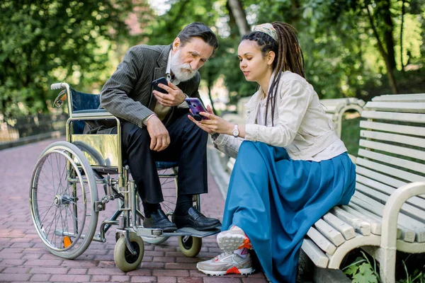 Ein älterer Patient sitzt im Rollstuhl, spricht mit seiner Pflegerin, Krankenschwester oder Enkelin, sitzt auf der Parkbank, schaut auf ihre Smartphones, lächelt und hat Spaß — Stockfoto