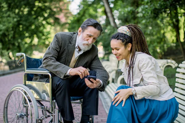 Porträt einer lächelnden jungen Hipsterfrau mit langen Dreadlocks, die auf der Parkbank sitzt, die Zeit mit ihrem behinderten Großvater im Rollstuhl genießt und Fotos auf dem Smartphone zeigt — Stockfoto