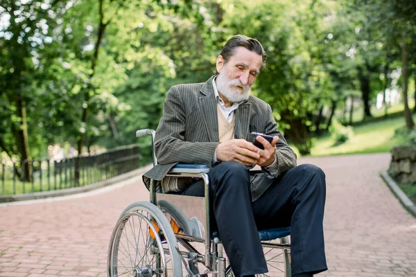Γενειοφόρος Καυκάσιος γέρος κάθεται μόνος του σε μια αναπηρική καρέκλα στο πάρκο, χρησιμοποιώντας το smartphone του για σέρφινγκ στο διαδίκτυο ή στα δίκτυα Σόκαϊλ. — Φωτογραφία Αρχείου