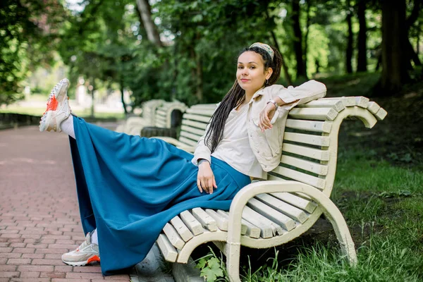 Outdoors portrait of modern hipster girl with dreadlocks wearing blue skirt and white t-shirt, sitting on the bench in a city park with one leg raised and having fun