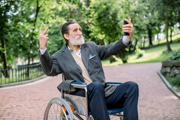 Glücklicher älterer behinderter oder bärtiger Mann, der im Rollstuhl im Park sitzt und sich mit seinem Smartphone vor dem Hintergrund der Parklandschaft fotografiert — Stockfoto