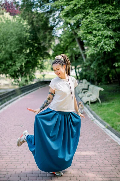 Retrato completo de menina bonita com dreadlocks dançando no parque da cidade, olhando para sua saia longa azul e sorrindo — Fotografia de Stock