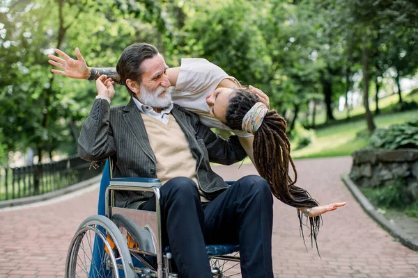 Porträt eines gutaussehenden bärtigen älteren Mannes, der im Rollstuhl in einem Stadtpark sitzt und sich freut, seine kreative Pflegerin oder Enkelin hinter sich zu sehen, die große Überraschung macht — Stockfoto