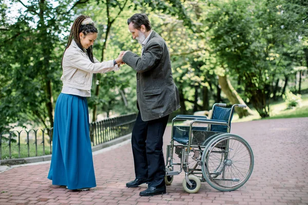 Älterer behinderter Mann, der versucht, sich mit Hilfe seiner hübschen Enkelin mit langen Dreadlockhaaren aus dem Rollstuhl zu erheben, lächelt und erfreut sich guter Gesundheit nach einer Krankheit — Stockfoto