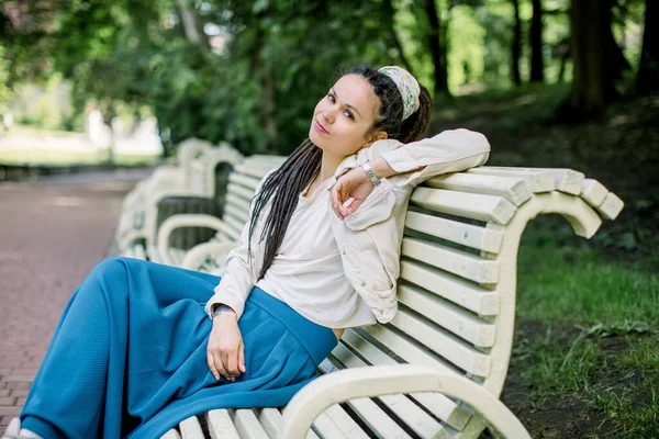 Contentada menina caucasiana feliz no desgaste casual, com dreadlocks em sua cabeça está descansando no banco contra fundo do belo parque da cidade — Fotografia de Stock