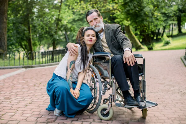 Junge Sozialarbeiterin oder Enkelin kümmert sich beim Spazierengehen im Park um einen älteren Mann im Rollstuhl, lächelt und umarmt sich, blickt in die Kamera. Verschiedene Generationen von Menschen — Stockfoto