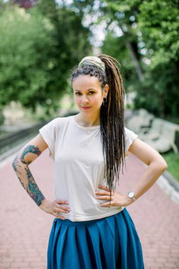 Rastalı, dövmeli, beyaz tişörtlü, mavi etekli çekici hippi kız portresi. Sabahları şehir parkında kameraya poz veriyor.