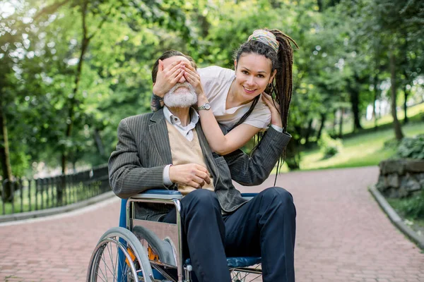 Junge lächelnde Kaukasierin mit Dreadlockhaaren, die die Augen ihres älteren behinderten Großvaters im Rollstuhl bedeckt und ihn bei einem Spaziergang im Park überrascht — Stockfoto