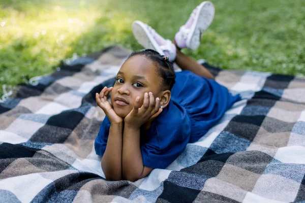 Happy smiling African girl child lying on blanket in summer park, leaning her face on her hands and looking at camera. Happy African child resting on green grass in park
