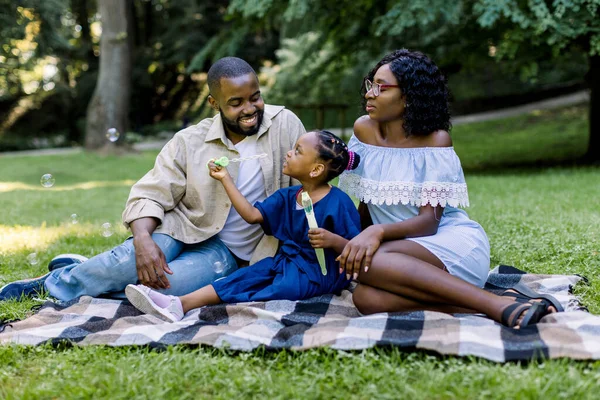 Feliz joven familia africana jugando con burbujas de jabón con su linda hija en el parque al aire libre en verano o primavera, sentada en la manta. Concepto de día familiar — Foto de Stock