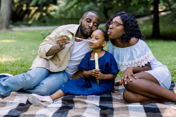 Tiempo en familia, vacaciones, ocio juntos. Alegre afro-americano famile con niña soplando burbujas juntos, divirtiéndose en un picnic en el parque de la ciudad, sentado en manta a cuadros — Foto de Stock