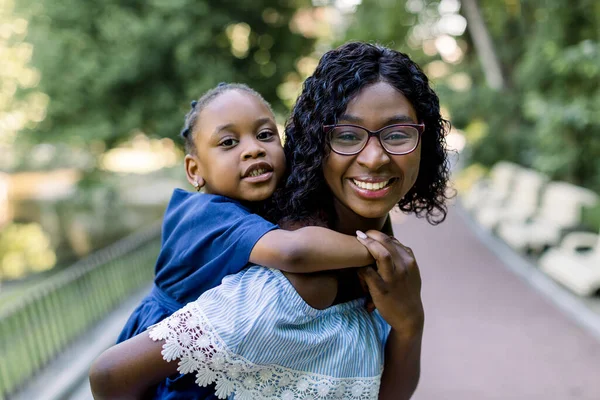 彼女の魅力的な笑顔の母親と一緒にピギーバックに乗ってかわいいアフリカの女の子の娘は、緑の木々と都市公園を歩いています。閉じる上のポルノの幸せな若いです母とともに娘piggybacking — ストック写真