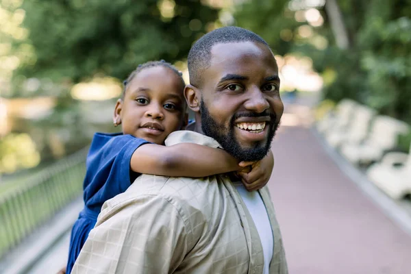 ハンサムなアフリカ系アメリカ人の父親は、彼のかわいい娘を屋外の都市公園に運んでいます。パパ運ぶ彼女の娘オンピギーバックでザ性質. — ストック写真