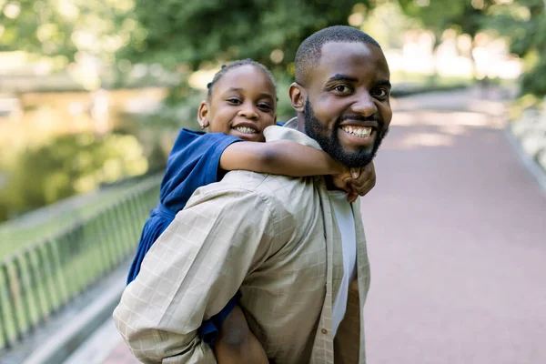 公園で遊んで、ハンサムな若い暗い肌の父と彼の笑いの小さな娘。アフリカ人の父親は自然の中で外に彼の娘をピギーバック。肖像画を閉じる — ストック写真