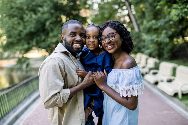 Sonriente retrato familiar alegre en el parque. Retrato de la joven familia Arican, de pie juntos en el fondo del callejón del parque, sosteniendo a su pequeña hija en las manos y riendo a la cámara — Foto de Stock