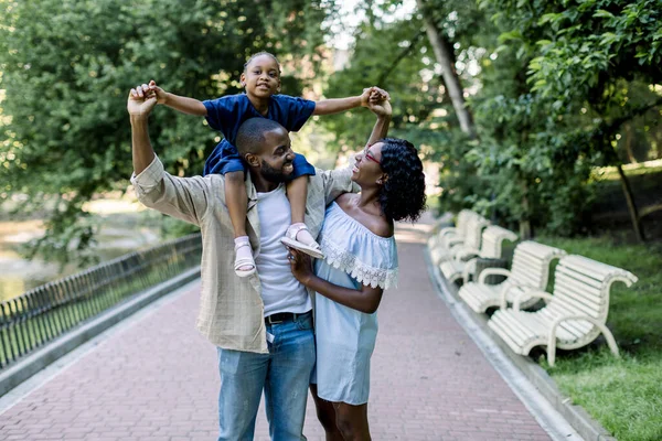 Família na natureza no parque. Bonito homem afro-americano pai carregando sua filhinha nos ombros, olhando para sua esposa muito sorridente e curtindo andar ao ar livre — Fotografia de Stock
