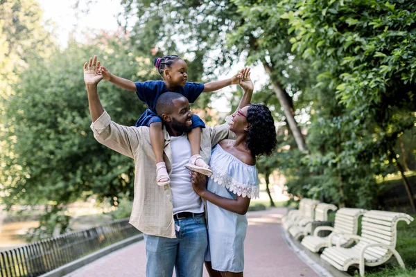 Mladý šťastný rozesmátý africký pár rodiče, trávit čas spolu a užívat si teplý letní večer v parku, procházky se svou malou rozkošnou dceruškou, sedí na ramenou otce — Stock fotografie