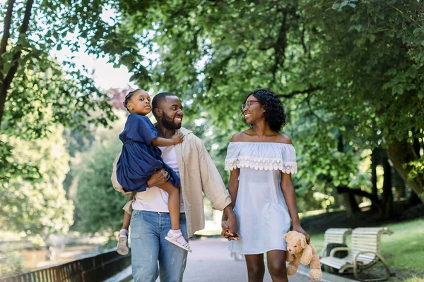 Familia africana joven feliz pasar tiempo al aire libre en el parque en un día de verano. alegre sonriente pareja cogida de la mano y caminando por el callejón del parque, hombre sostiene linda hija pequeña en vestido azul — Foto de Stock