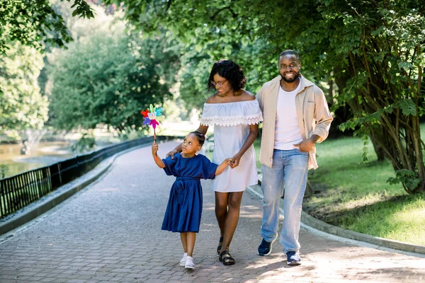 Joven familia afroamericana, padre, madre y niña, jugando colorido juguete molino de viento en el parque al aire libre, disfrutar de divertido tiempo en familia con la luz del sol por la mañana o por la noche y árboles verdes en el fondo . — Foto de Stock