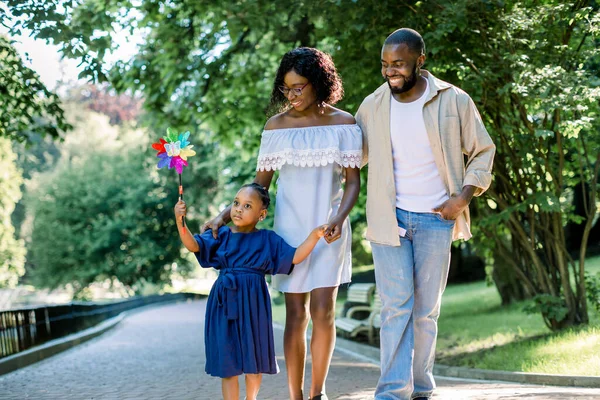 Família feliz, tempo alegre juntos ao ar livre. Menina de pele escura em vestido azul, soprando brinquedo colorido moinho de vento, enquanto caminhava no beco do parque com seus jovens pais felizes no dia de verão — Fotografia de Stock