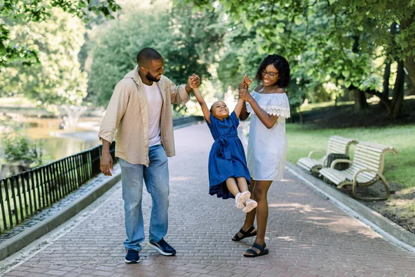 Feliz familia africana en el parque en la tarde soleada de verano. Mamá, papá y feliz hija caminan al atardecer, padres sosteniendo las manos de una niña, volando en el aire. El concepto de una familia feliz . — Foto de Stock