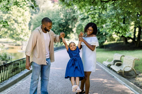 Feliz familia afroamericana disfrutando de la actividad al aire libre en el parque de la ciudad, tomados de la mano, caminando. Riéndose niña en vestido azul, toma las manos de su mamá y su papá y vuela en el aire — Foto de Stock