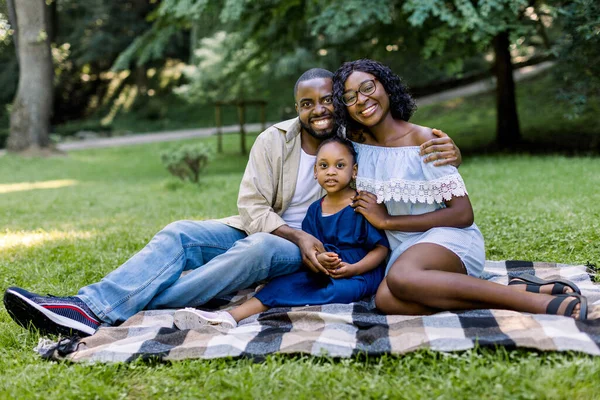 Tempo de ligação familiar. Feliz jovem família africana de três sorrindo enquanto sentado em elegante cobertor quadriculado no parque, com grama verde e árvores no fundo — Fotografia de Stock