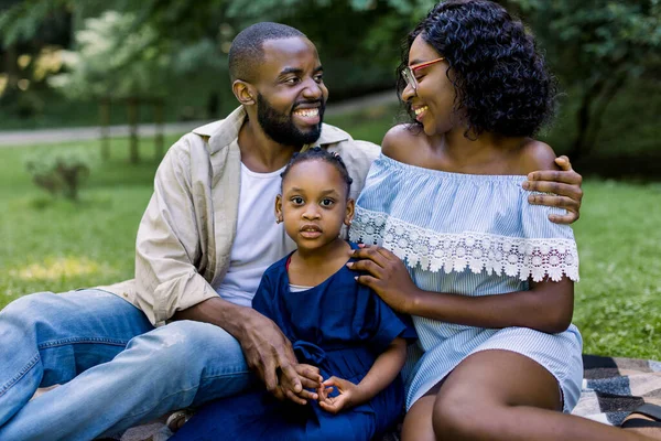 Joven familia africana feliz con su hija pequeña pasar su tiempo en el parque en el picnic, sentado en la hierba verde y sonriendo mirando el uno al otro. Familia feliz en el amor concepto — Foto de Stock