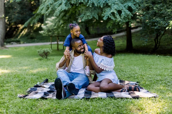 Feliz familia africana de tres teniendo un picnic en el parque en el día de verano. Familia joven al aire libre en un parque, sentada en una manta a cuadros. Niña chica es piggybacking en su papá — Foto de Stock