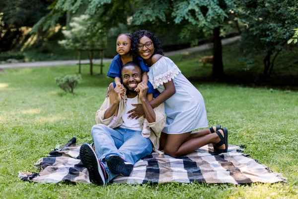 Um piquenique familiar no parque. Família africana feliz com menina, vestindo roupas casuais e elegantes, sentado em xadrez xadrez no parque e sorrindo para a câmera. Conceito de família feliz . — Fotografia de Stock