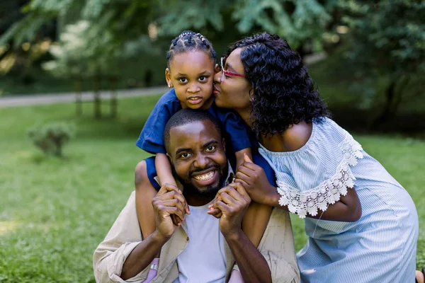Familia africana feliz con niña en un picnic en el parque. Sonriente padre guapo sostiene a su linda hija en los hombros, la madre bonita besa a la niña. Familia feliz — Foto de Stock