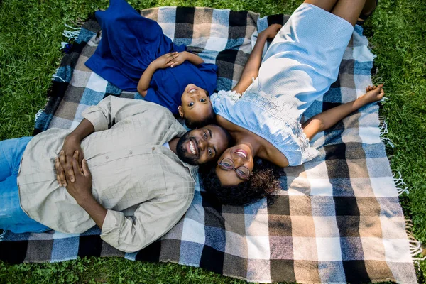 Happy African family, mom, dad and cute daughter lie and play on the checkered blanket in the park on a sunny summer day. The concept of family happiness. The view from the top