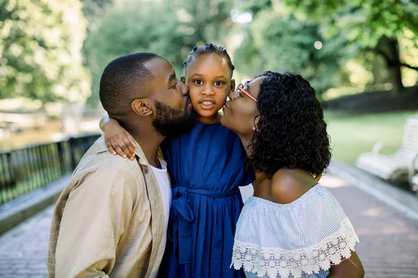 Adorable retrato de verano de la joven familia africana de tres posando juntos al aire libre en un hermoso parque. Los padres encantadores están besando a su hija pequeña con vestido azul. De cerca. — Foto de Stock