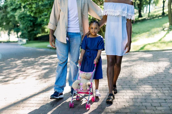 街の公園で彼女の若い両親と一緒に歩いている間、ベビーカーのおもちゃとテディベアで遊んで青いドレスを着た愛らしい小さなアフリカの女の子のトリミングされたイメージ。幸せな子供時代と家族 — ストック写真