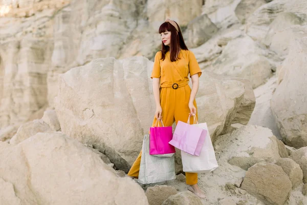 Horizontal view of young female model in stylish clothes, holding full colorful shopping bags, posing in front of sand canyon or quarry outdoors. Shopping concept, fashion model. Copy space