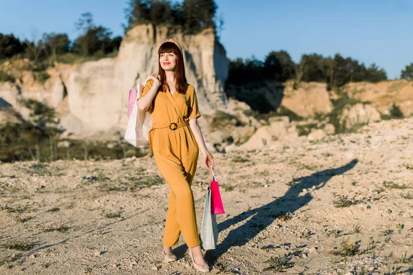 Retrato de comprimento total de bela mulher de moda de cabelos retos em macacões amarelos, de pé com sacos de compras coloridos nas mãos na frente da pedreira de areia e desfrutando do pôr do sol de verão — Fotografia de Stock