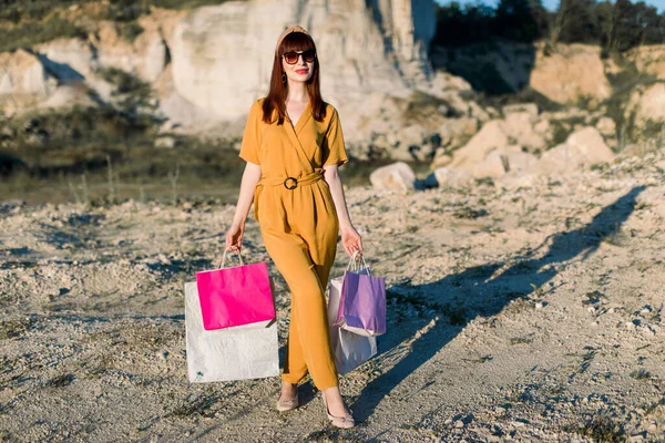 Full length portrait of a young red haired girl in sunglasses and fashionable yellow overalls, carrying shopping bags while walking in sand quarry. Horizontal view. Summer shopping sale