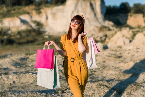 Photo of pretty happy cute woman, wearing yellow overalls, with colorful shopping bags in hands, enjoying of new purchases over summer sunset in sand quarry background. Fashion and style lifestyle — Stock Photo, Image