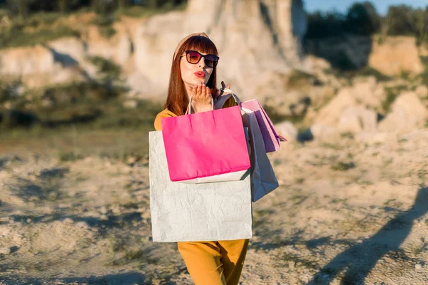 Summer bright portrait of beautiful young woman in yellow clothes and sunglasses, making air kiss to camera, while holding colorful shopping bags, posing on the background of sand quarry — Stock Photo, Image
