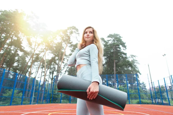 Beautiful young blond girl in sports clothing holding yoga mat, while walking before sport training outdoors, on professional training court. Sport and fitness concept, summer workout