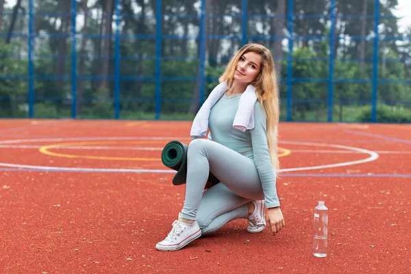 Attraktiv ung blond fitness kvinna bär grå sportkläder poserar utomhus på sport domstol med rött golv med en träningsmatta, ler mot kameran. Sport och träning utomhus — Stockfoto