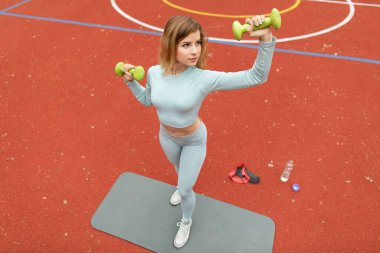 Spor figürlü çekici bir genç kız, spor eğitmeni, yeşil halterlerle egzersiz yapıyor, gri formda, açık havada kırmızı kaplamalı spor kortunda duruyor. Üst Manzara. Açık havada egzersiz.