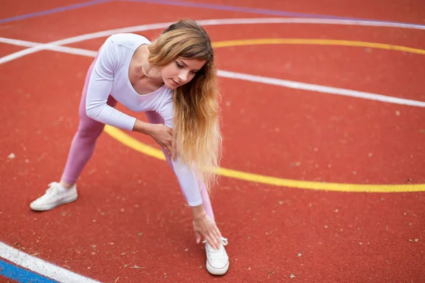 Νεαρή όμορφη ξανθιά αθλήτρια με ροζ αθλητικά ρούχα, στέκεται στο κόκκινο επιχρισμένο γήπεδο, τεντώνεται και ετοιμάζεται να τρέξει. Αθλητισμός και τρέξιμο στην ύπαιθρο. Αντιγραφή χώρου — Φωτογραφία Αρχείου