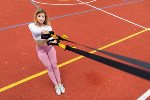 Pretty young Caucasian girl in pink sportswear exercising with suspension trainer sling, doing squats, outdoors at red sport court. Fitness concept with trx trainer