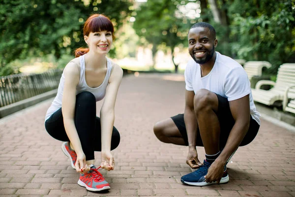 Casal de corredores, cara africano e menina caucasiana, amarrando atacadores de tênis de corrida no parque, sorrindo e olhando para a câmera. Estilo de vida saudável, motivação jogging, pessoas saudáveis felizes — Fotografia de Stock