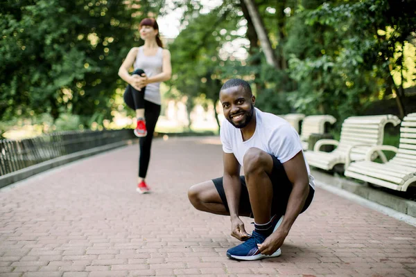 Bonito sorrindo homem africano atando seus sapatos e se preparando para correr e esporte ao ar livre no parque da cidade com sua namorada, mulher muito caucasiana, de pé no fundo e alongamento — Fotografia de Stock