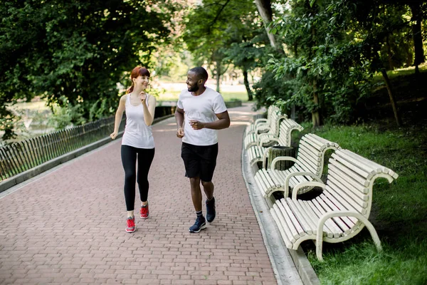 Horizontal tiro de jovem casal multi racial, vestindo camisetas brancas e calças pretas, correndo no beco do parque da cidade, no início da manhã com o nascer do sol no fundo — Fotografia de Stock